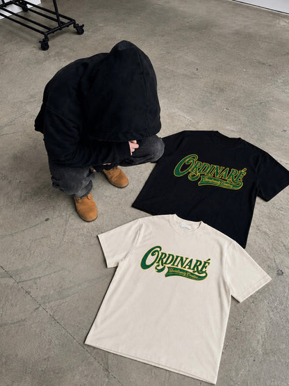 Two t-shirts with a green 'Ordinaire' graphic logo are displayed on a concrete floor, one in cream and one in black, with a person in a black hoodie crouching nearby.